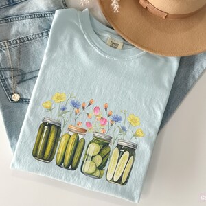 Puede incluir: Una camiseta azul claro con un gr&aacute;fico de cuatro frascos de vidrio llenos de pepinillos y cubiertos con una variedad de flores. El texto "COMFORT COLORS" est&aacute; impreso en la camiseta.