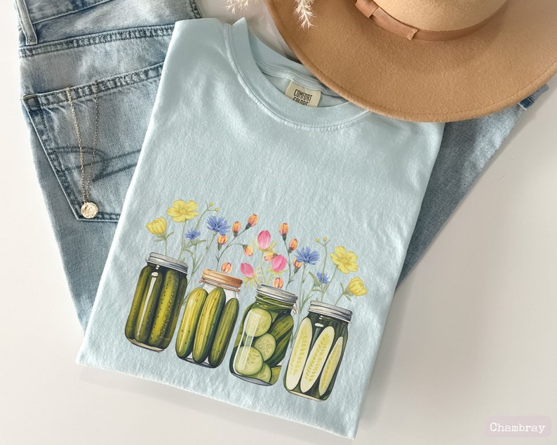 Puede incluir: Una camiseta azul claro con un gr&aacute;fico de cuatro frascos de vidrio llenos de pepinillos y cubiertos con una variedad de flores. El texto "COMFORT COLORS" est&aacute; impreso en la camiseta.