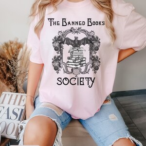 May include: A pink t-shirt with a black graphic design that says "The Banned Books Society". The graphic features a bat flying over a stack of books with a decorative frame.