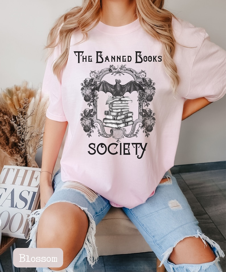 May include: A pink t-shirt with a black graphic design that says "The Banned Books Society". The graphic features a bat flying over a stack of books with a decorative frame.