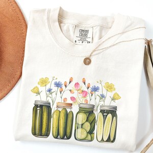 Puede incluir: Una camiseta de color crema con un gr&aacute;fico de cuatro tarros de pepinillos con eneldo verde y flores amarillas. El texto "COMFORT COLORS" est&aacute; impreso en la camiseta.