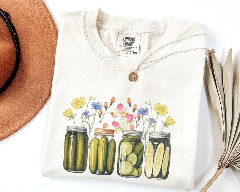 Puede incluir: Una camiseta de color crema con un gr&aacute;fico de cuatro tarros de pepinillos con eneldo verde y flores amarillas. El texto "COMFORT COLORS" est&aacute; impreso en la camiseta.