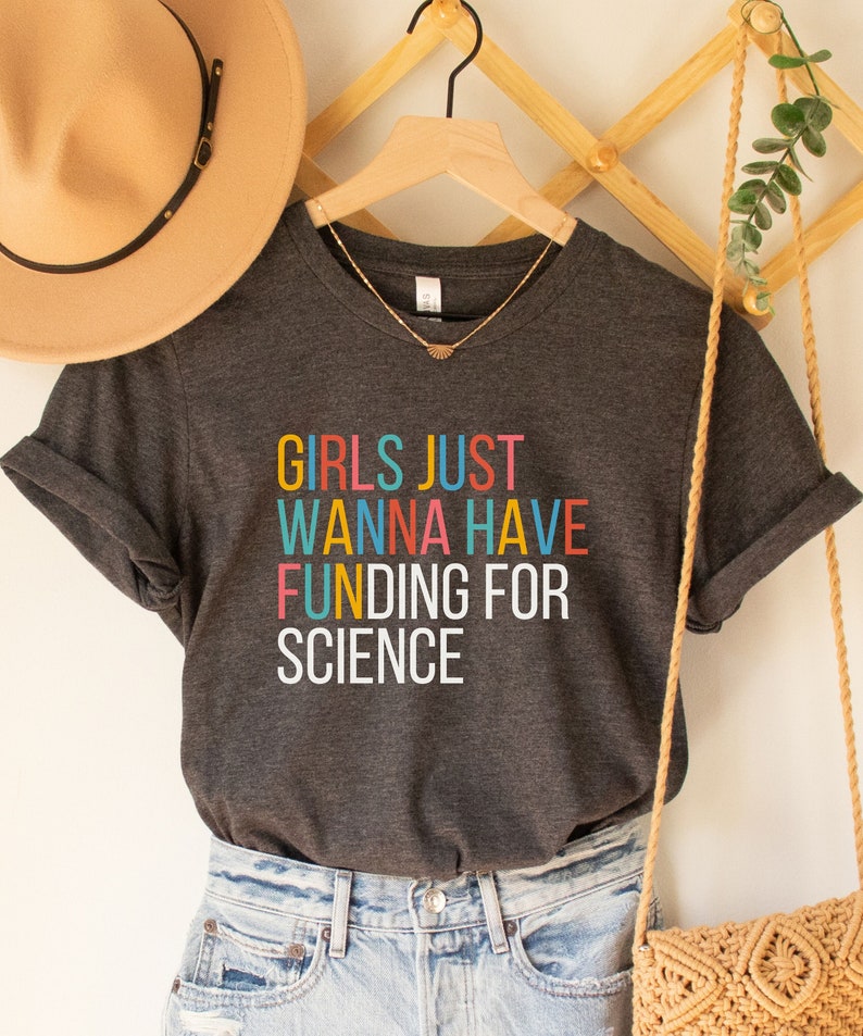 以下が含まれることがあります： カラフルな文字で「GIRLS JUST WANNA HAVE FUNDING FOR SCIENCE」と書かれたダークグレーのTシャツです。