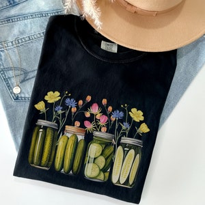 Puede incluir: Camiseta negra con un gr&aacute;fico de cuatro tarros de vidrio llenos de pepinillos y cubiertos de flores silvestres. Las flores son amarillas, rosas, azules y verdes.