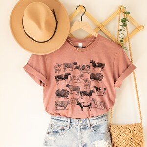 May include: A heather mauve t-shirt with a vintage illustration of various breeds of cows. The shirt is hanging on a wooden hanger with a brown hat and a woven bag.