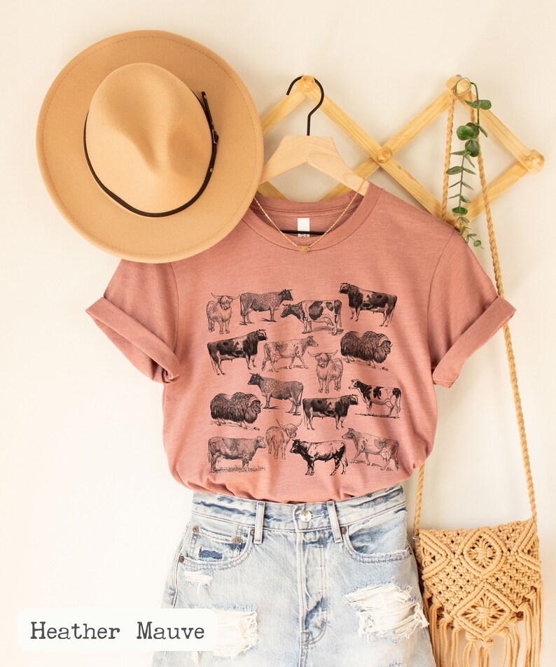 May include: A heather mauve t-shirt with a vintage illustration of various breeds of cows. The shirt is hanging on a wooden hanger with a brown hat and a woven bag.
