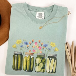 Puede incluir: Una camiseta azul claro con un dise&ntilde;o floral y cuatro tarros de pepinillos. Los tarros est&aacute;n llenos de pepinillos verdes y tienen un dise&ntilde;o floral en las tapas. El texto "COMFORT COLORS" est&aacute; impreso en la etiqueta.