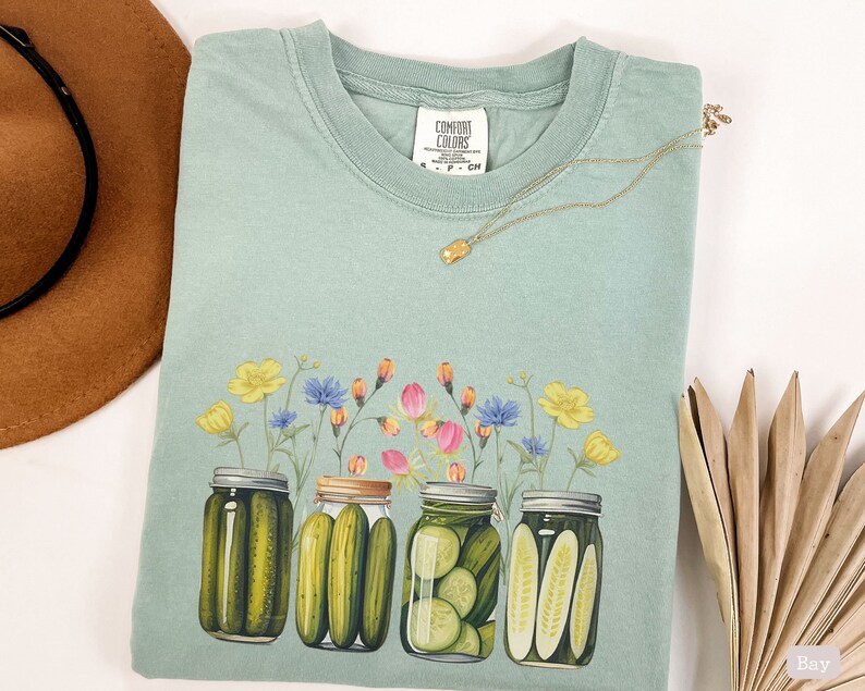 Puede incluir: Una camiseta azul claro con un dise&ntilde;o floral y cuatro tarros de pepinillos. Los tarros est&aacute;n llenos de pepinillos verdes y tienen un dise&ntilde;o floral en las tapas. El texto "COMFORT COLORS" est&aacute; impreso en la etiqueta.