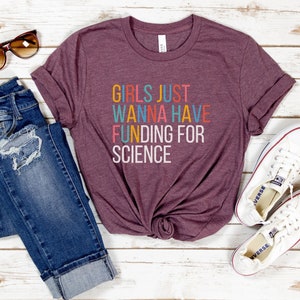 以下が含まれることがあります： カラフルな文字で「Girls Just Wanna Have Funding For Science」と書かれた、マルーンのTシャツです。