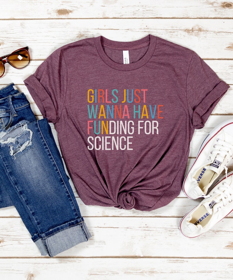 以下が含まれることがあります： カラフルな文字で「Girls Just Wanna Have Funding For Science」と書かれた、マルーンのTシャツです。