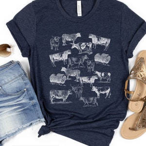 May include: A heather navy blue t-shirt with a white graphic print of various cow breeds. The shirt has a crew neck and short sleeves.