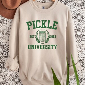 May include: A beige sweatshirt with a green graphic that says "Pickle University" with a pickle jar and the text "Est. 1893".
