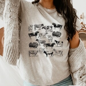 May include: A gray t-shirt with a black and white print of various cows and bulls. The shirt has a crew neck and short sleeves. The word "Ash" is written in white on a black background in the lower right corner of the image.