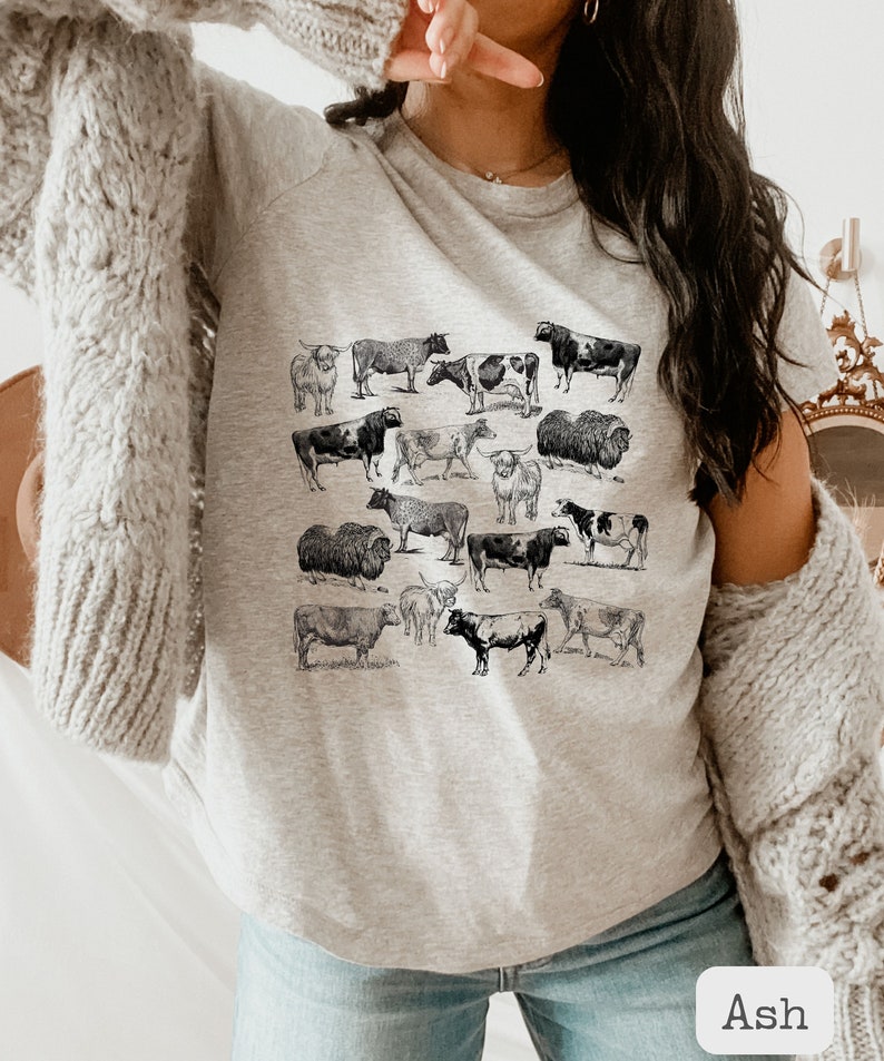 May include: A gray t-shirt with a black and white print of various cows and bulls. The shirt has a crew neck and short sleeves. The word "Ash" is written in white on a black background in the lower right corner of the image.