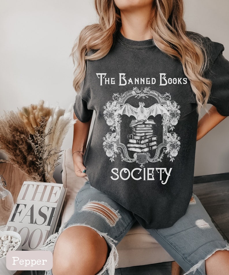 May include: A black t-shirt with a white graphic design featuring a bat and a stack of books. The text "The Banned Books Society" is printed on the shirt.