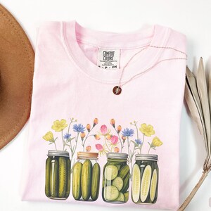 Puede incluir: Camiseta rosa con un gr&aacute;fico de cuatro tarros de pepinillos con flores que salen de la parte superior. Los tarros est&aacute;n llenos de pepinillos verdes y las flores son amarillas, rosas y azules. La camiseta tiene una etiqueta que dice "Comfort Colors".