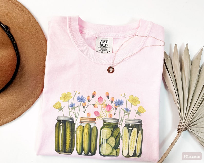 Puede incluir: Camiseta rosa con un gr&aacute;fico de cuatro tarros de pepinillos con flores que salen de la parte superior. Los tarros est&aacute;n llenos de pepinillos verdes y las flores son amarillas, rosas y azules. La camiseta tiene una etiqueta que dice "Comfort Colors".