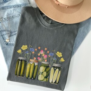 Puede incluir: Una camiseta gris oscura con un gr&aacute;fico de cuatro tarros de cristal llenos de pepinillos y coronados con flores silvestres. Las flores son amarillas, rosas y azules.