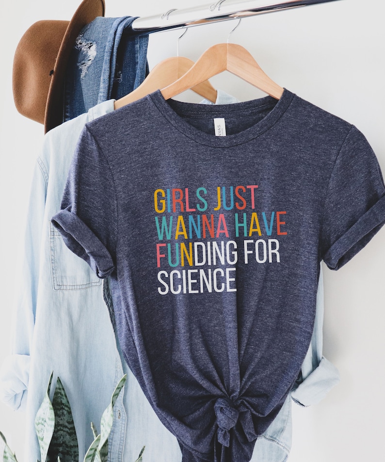 以下が含まれることがあります： 虹色の文字で「Girls Just Wanna Have Funding For Science」と書かれた濃い青色のTシャツです。