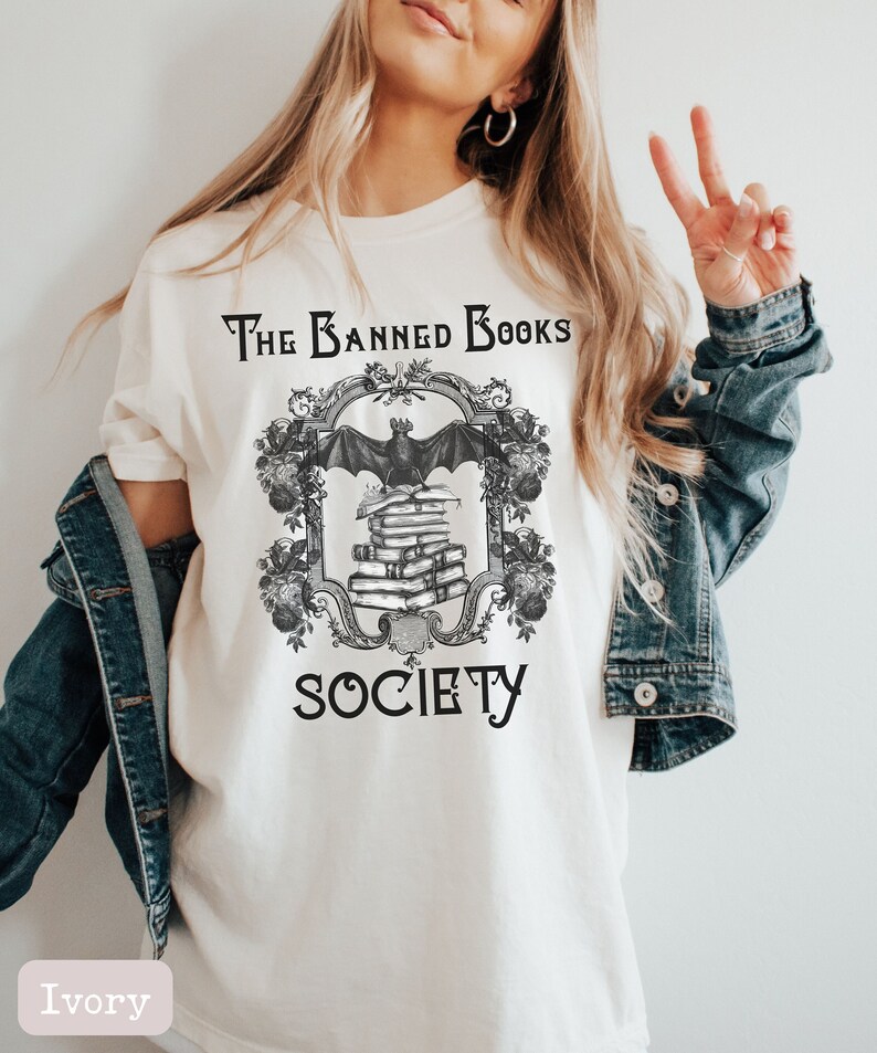 May include: A white t-shirt with a black and white graphic design featuring a bat perched on a stack of books. The graphic is framed with a decorative border and the text "The Banned Books Society".