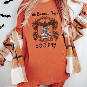 May include: A burnt orange t-shirt with a graphic design of a bat perched on a stack of books. The graphic is framed with a decorative border and the text "The Banned Books Society".