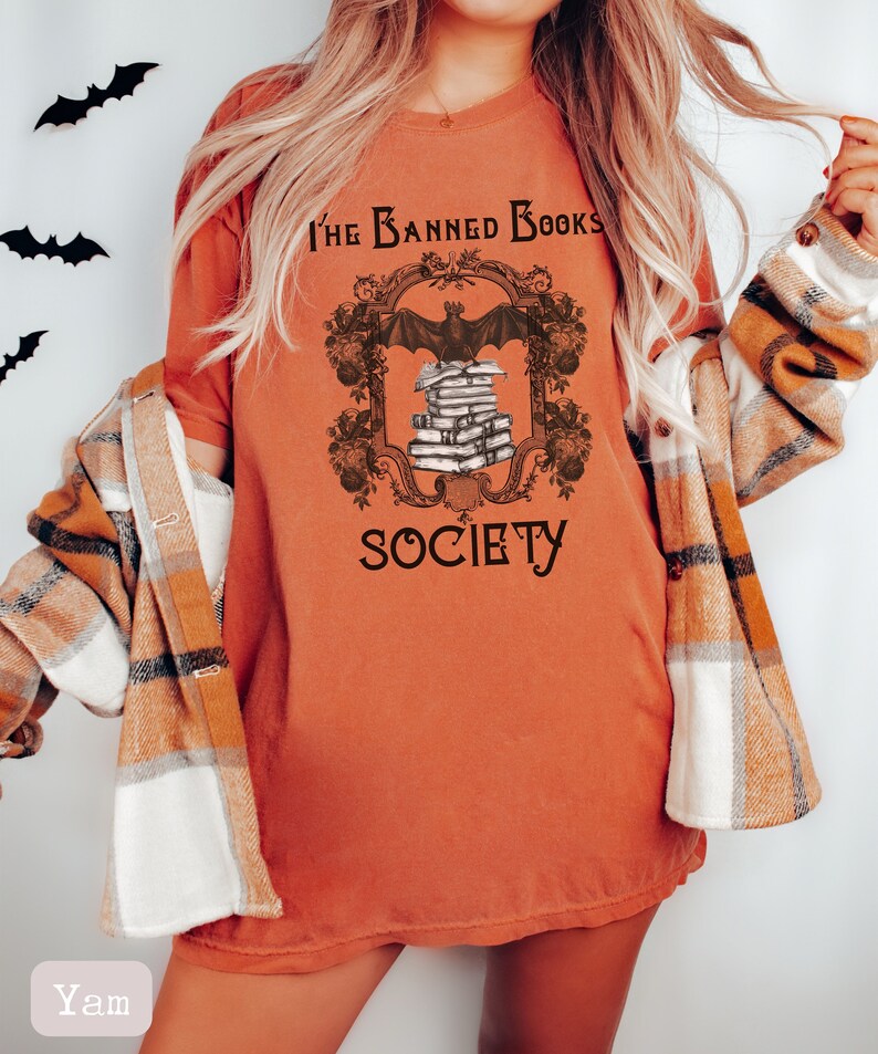 May include: A burnt orange t-shirt with a graphic design of a bat perched on a stack of books. The graphic is framed with a decorative border and the text "The Banned Books Society".