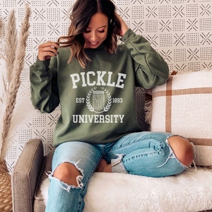 May include: A green sweatshirt with the text "Pickle University" in white, featuring a white illustration of a jar of pickles and the text "Est. 1893".
