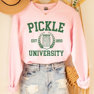 May include: Pink sweatshirt with the text "PICKLE UNIVERSITY" in green letters. The text is surrounded by a green wreath with a jar of pickles in the centre. The text "EST 1893" is below the wreath.