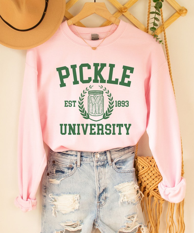 May include: Pink sweatshirt with the text "PICKLE UNIVERSITY" in green letters. The text is surrounded by a green wreath with a jar of pickles in the centre. The text "EST 1893" is below the wreath.