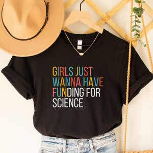 以下が含まれることがあります： カラフルな文字で「Girls Just Wanna Have Funding For Science」と書かれた黒いTシャツです。