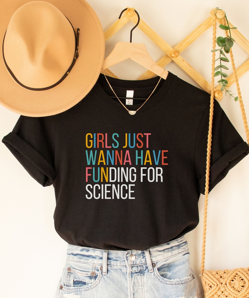 以下が含まれることがあります： カラフルな文字で「Girls Just Wanna Have Funding For Science」と書かれた黒いTシャツです。