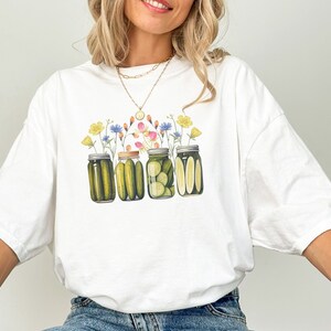 Puede incluir: Camiseta blanca con un gr&aacute;fico de cuatro tarros de pepinillos con flores en la parte superior. Los tarros est&aacute;n llenos de pepinillos verdes y las flores son rosas, amarillas, azules y blancas.