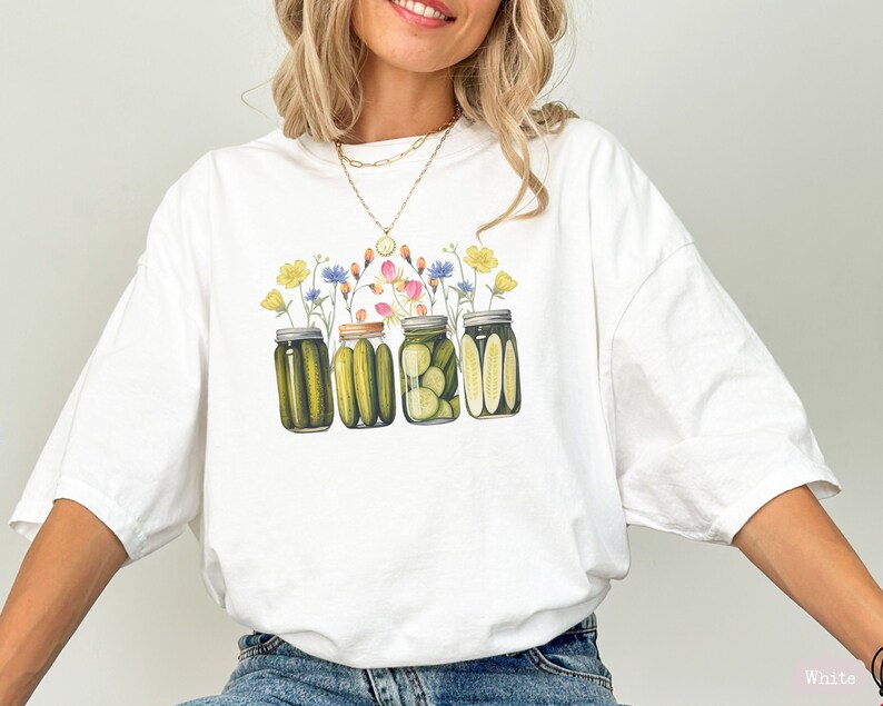 Puede incluir: Camiseta blanca con un gr&aacute;fico de cuatro tarros de pepinillos con flores en la parte superior. Los tarros est&aacute;n llenos de pepinillos verdes y las flores son rosas, amarillas, azules y blancas.