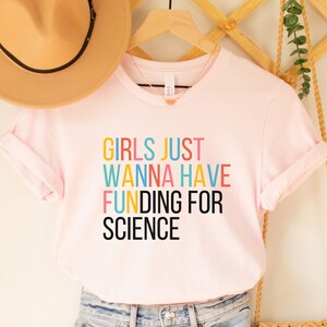以下が含まれることがあります： ピンクのTシャツに「Girls Just Wanna Have Funding For Science」というカラフルな文字が書かれています。