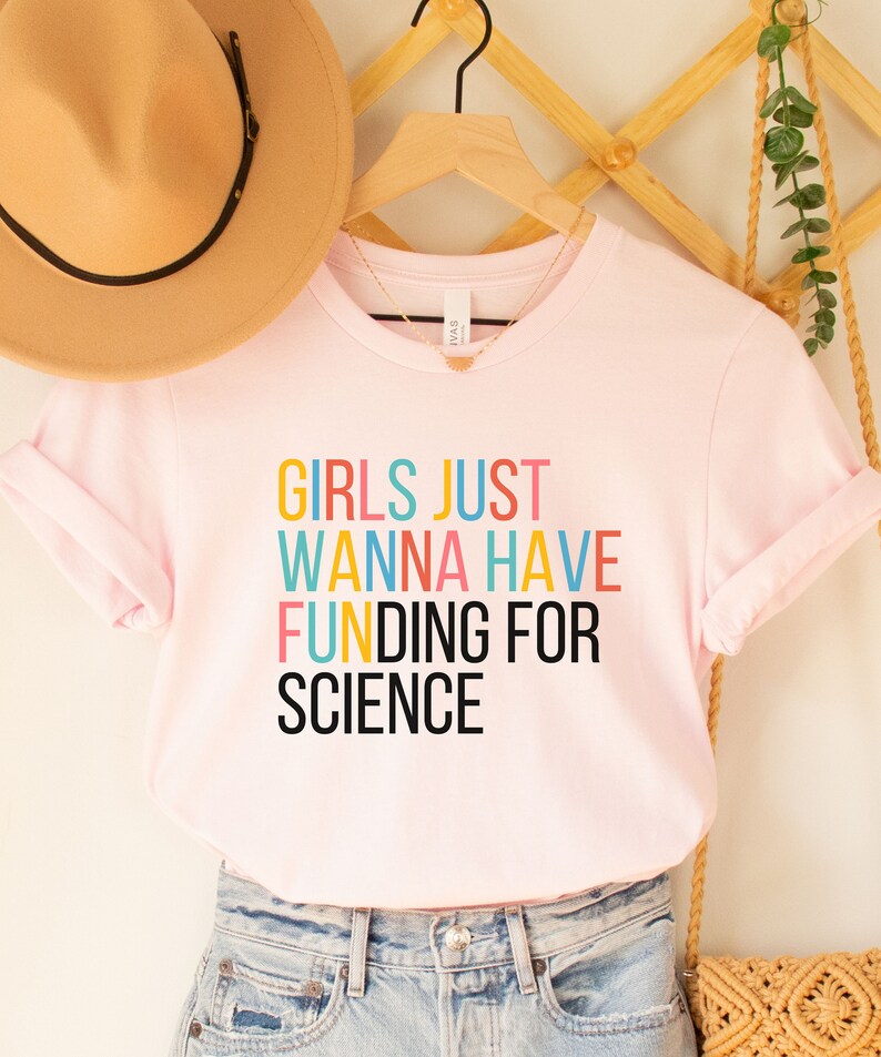 以下が含まれることがあります： ピンクのTシャツに「Girls Just Wanna Have Funding For Science」というカラフルな文字が書かれています。
