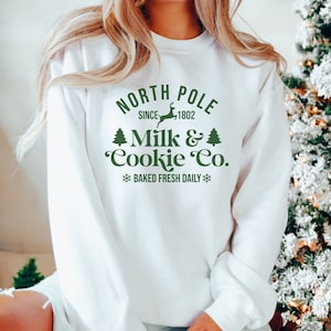 May include: White sweatshirt with a green graphic that reads "NORTH POLE SINCE 1802 MILK & COOKIE CO." with a reindeer and Christmas tree illustrations. The text "BAKED FRESH DAILY" is also included. The sweatshirt is a festive holiday apparel item.
