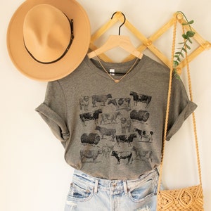 May include: A gray t-shirt with a vintage illustration of cows and sheep. The shirt is hanging on a wooden hanger with a brown hat and a straw bag.