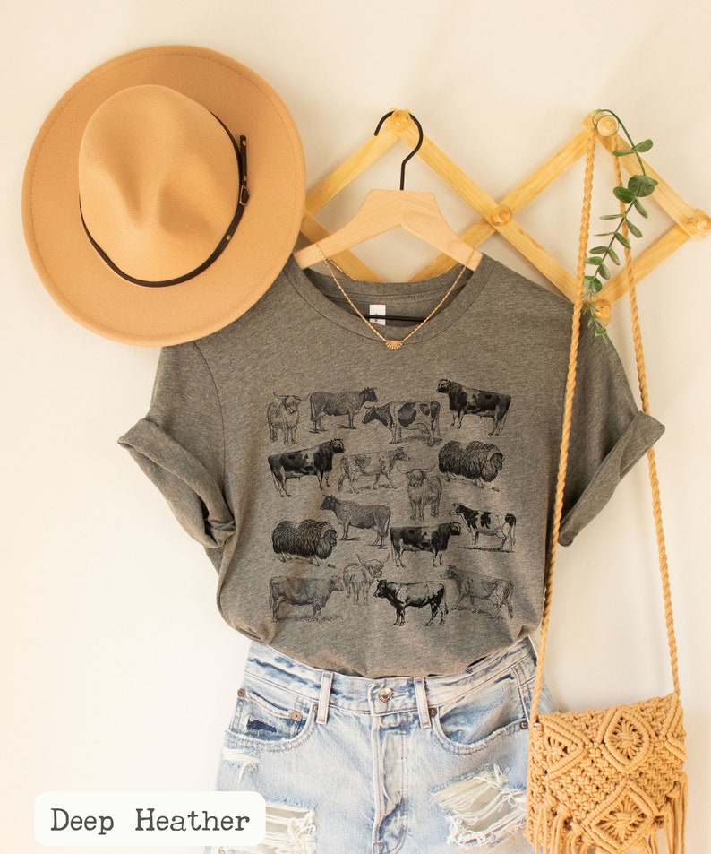 May include: A gray t-shirt with a vintage illustration of cows and sheep. The shirt is hanging on a wooden hanger with a brown hat and a straw bag.