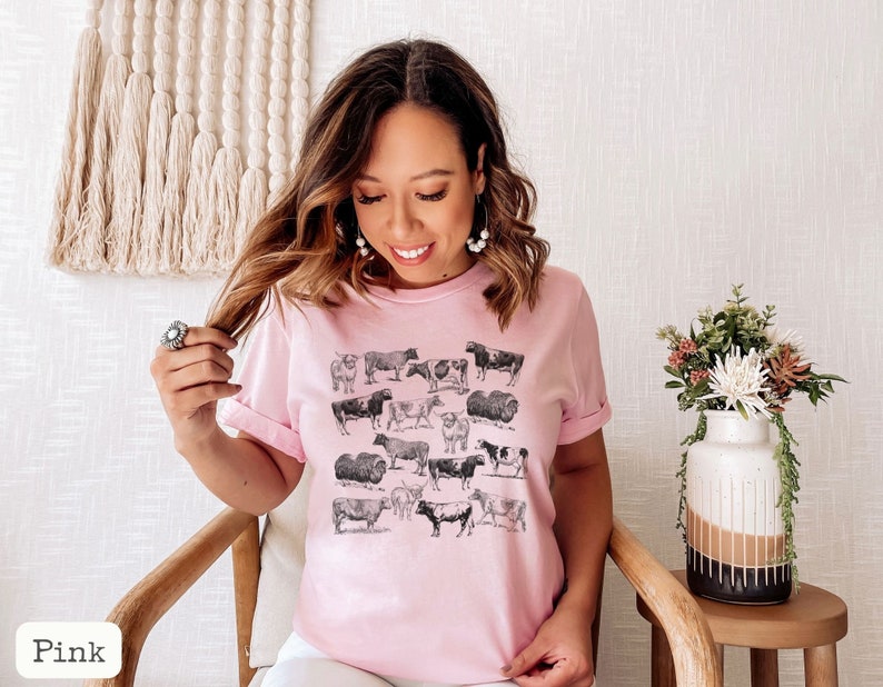 May include: A pink t-shirt with a black and white illustration of cows. The shirt has a crew neck and short sleeves.