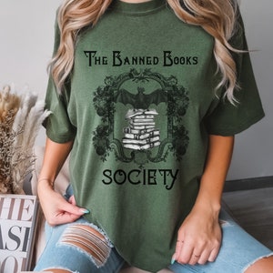 May include: A green t-shirt with a black graphic of a bat flying over a stack of books. The graphic is framed with a black ornate design and the text "The Banned Books Society".