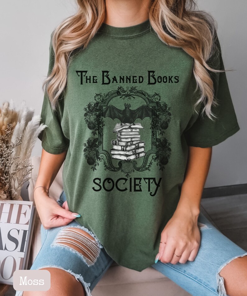 May include: A green t-shirt with a black graphic of a bat flying over a stack of books. The graphic is framed with a black ornate design and the text "The Banned Books Society".