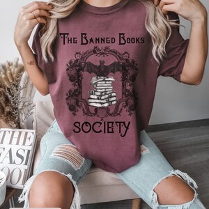 May include: A maroon t-shirt with a graphic of a bat perched on a stack of books. The graphic is framed with an ornate design and the text "The Banned Books Society".