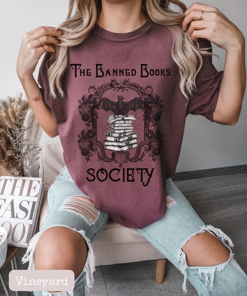 May include: A maroon t-shirt with a graphic of a bat perched on a stack of books. The graphic is framed with an ornate design and the text "The Banned Books Society".