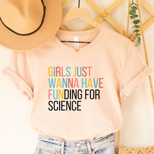 以下が含まれることがあります： 薄いピンクのTシャツに、カラフルな文字で「Girls Just Wanna Have Funding For Science」と書かれています。