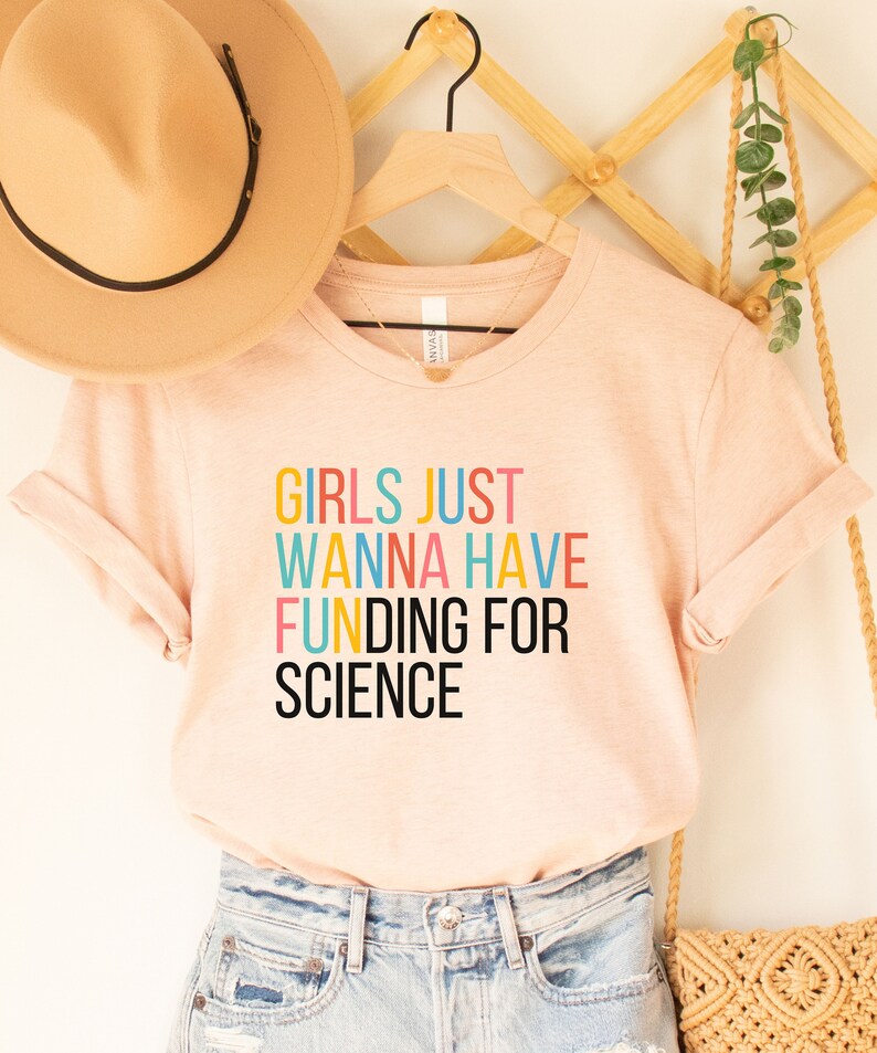 以下が含まれることがあります： 薄いピンクのTシャツに、カラフルな文字で「Girls Just Wanna Have Funding For Science」と書かれています。