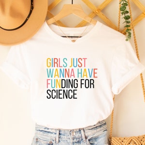 以下が含まれることがあります： カラフルな文字で「Girls Just Wanna Have Funding For Science」と書かれた白いTシャツです。