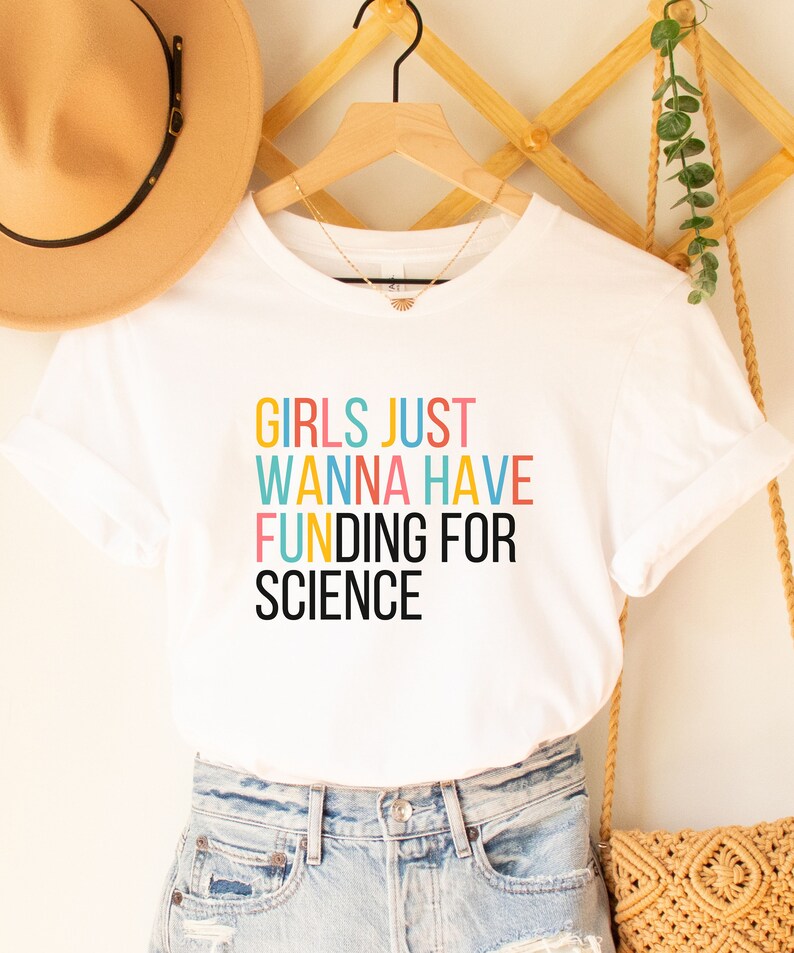 以下が含まれることがあります： カラフルな文字で「Girls Just Wanna Have Funding For Science」と書かれた白いTシャツです。