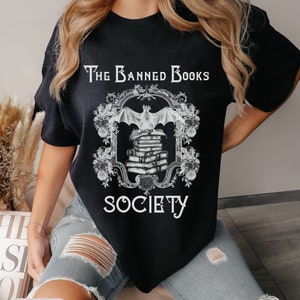 May include: Black t-shirt with a white graphic design of a bat flying over a stack of books. The graphic is framed with a decorative border and the text "The Banned Books Society".