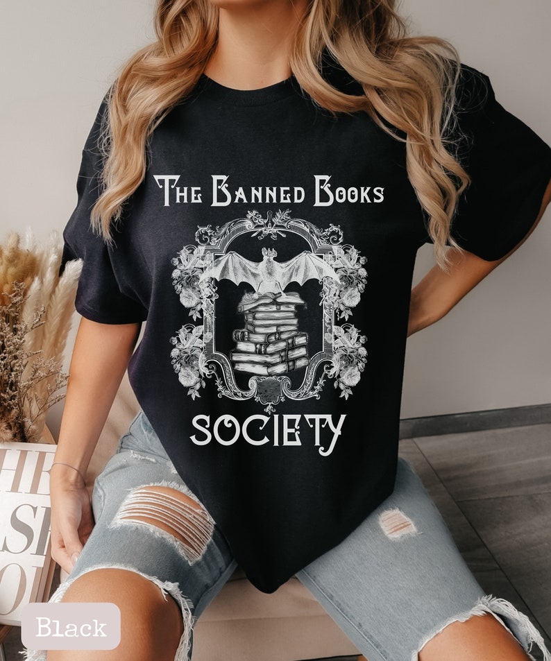 May include: Black t-shirt with a white graphic design of a bat flying over a stack of books. The graphic is framed with a decorative border and the text "The Banned Books Society".