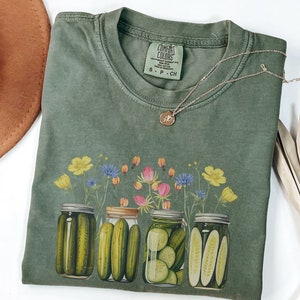 Puede incluir: Camiseta verde con un estampado de cuatro botes de pepinillos y flores silvestres. La etiqueta dice "COMFORT COLORS".